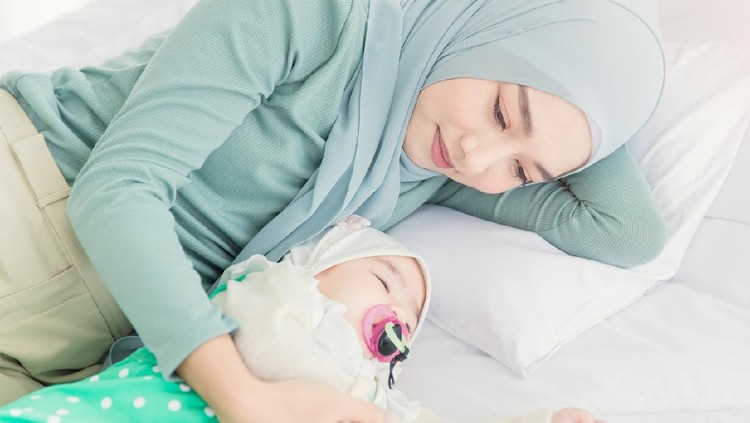 Happy muslim mother holding adorable little baby daughter wearing hijab in her arms on white bed in bedroom. Arab young mom wearing a head scarf, and she's sitting on the bed while holding her baby.