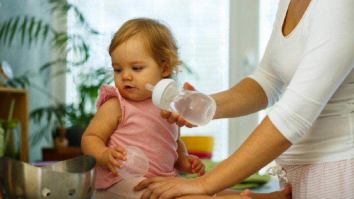 Shot of mid adult woman standing next to kitchen counter where her baby daughter is sitting and giving her bottle of water.