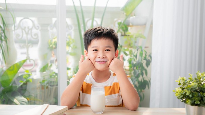Adorable happy young Asian boy having breakfast and drinking milk at home in the morning; looking at camera