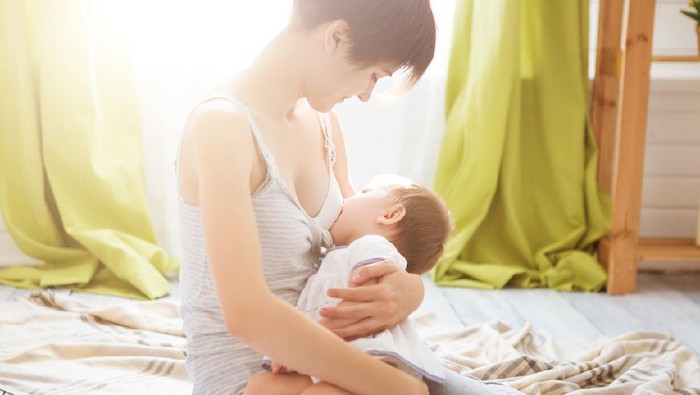 A portrait of a beautiful young Caucasian mother breastfeeding her adorable baby girl while working on a laptop in a kitchen at home.