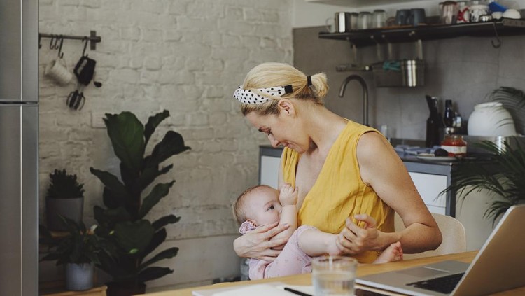 menyusui A portrait of a beautiful young Caucasian mother breastfeeding her adorable baby girl while working on a laptop in a kitchen at home.