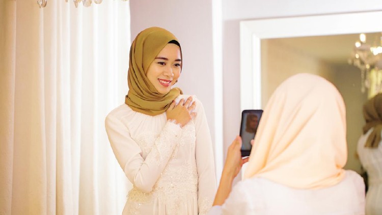 Young Muslim woman photographing her friend trying on a wedding dress in the bridal shop's fitting room.