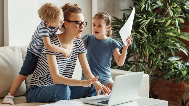 Tired young mother sitting on sofa and working with laptop and documents while little kids having fun and making noise