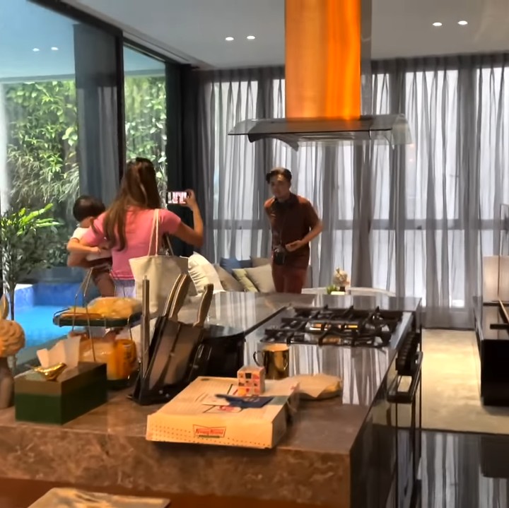 <p>Ketika masuk ke dalam rumah, tamu akan disambut dengan meja panjang di ruangan tengah yang dapat digunakan sebagai area tamu. Ada kolam renang yang dibatasi dengan jendela kaca untuk menambah suasana asri seperti berlbur di Bali. (Foto: YouTube NisNaz Channel)</p>