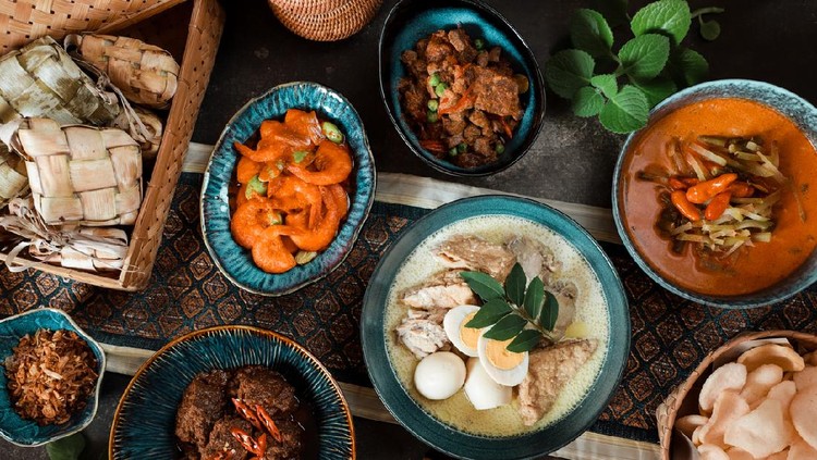 Ketupat Lebaran, the Indonesian elaborate meal of rice cake with meat and vegetable side dishes, served during Eid celebrations. The rice cake is cooked in diamond shaped casing made from woven coconut leaves. The accompanying dishes served are White Chicken Curry, Beef Rendang, Shrimps in Red Chili Paste, Spicy Liver Stew, Red Vegetable Curry Soup, and Shrimp Crackers. The dishes are arranged on a wooden table lined with a table liner. Several cumin leaves are used to decorate the table.