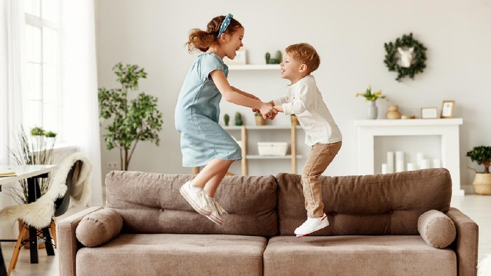 Side view of cheerful boy and girl holding hands and jumping on couch while having fun at home together