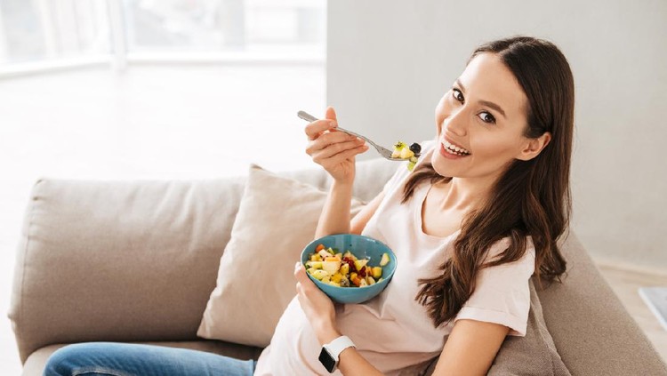 Pretty pregnant young woman having healthy breakfast while sitting on a couch