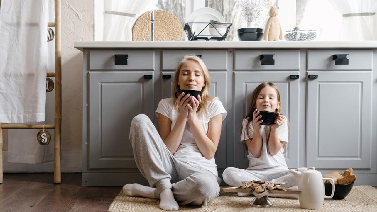 Dreamy mother sitting on kitchen floor together with her lovely daughter, enjoying morning coffee, drinking beverage and spending weekend in cozy apartment, resting at home