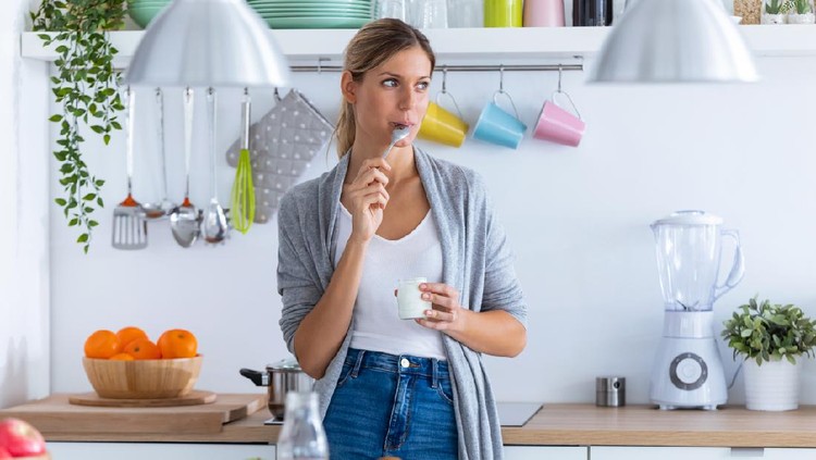 Shot of pretty young woman eating yogurt while standing in the kitchen at home.