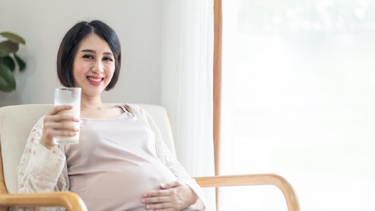 Young pretty Asian pregnant woman sits on armchair in front of white curtain holding milk and touching her tummy looks at camera with smile. Love care of pregnancy woman and baby health & food concept