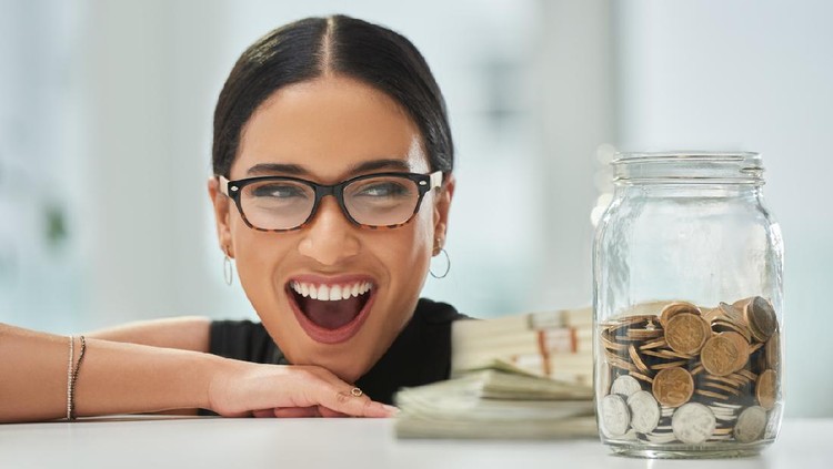 Cropped shot of an attractive young businesswoman looking excitedly over at a jar of coins and wads of cash in her office