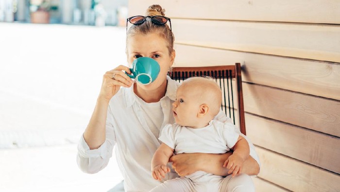 Caucasian young woman sits in a deck chair at the table, drinks coffee and holds the baby in her arms. Summer day in the city. Lifestyle portrait of mother with baby.