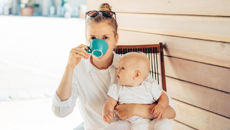 Caucasian young woman sits in a deck chair at the table, drinks coffee and holds the baby in her arms. Summer day in the city. Lifestyle portrait of mother with baby.