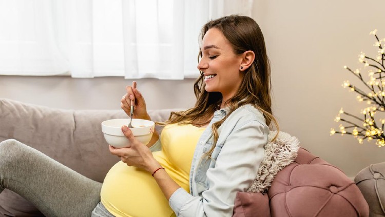 Beautiful young pregnant woman relaxing on sofa and eating corn flakes. Pregnancy concept.