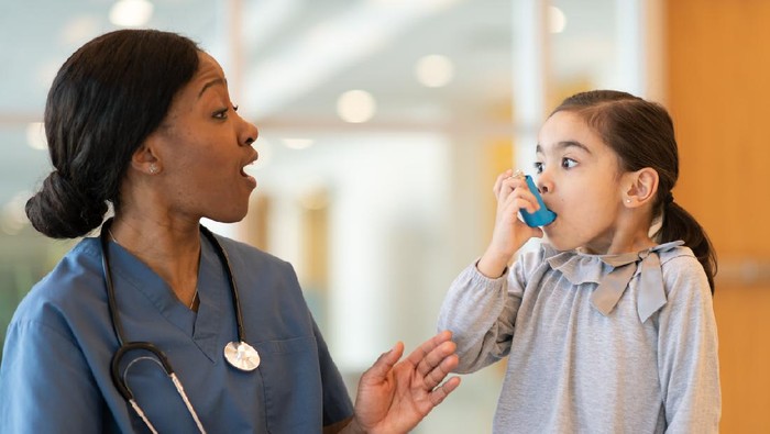 An ethnic girl with asthma is at a doctor's appointment. She is seated next to her African American doctor. The child is using a puffer. The doctor is watching and advising how to use the device properly.