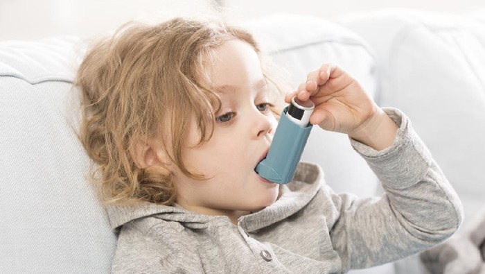An ethnic girl with asthma is at a doctor's appointment. She is seated next to her African American doctor. The child is using a puffer. The doctor is watching and advising how to use the device properly.