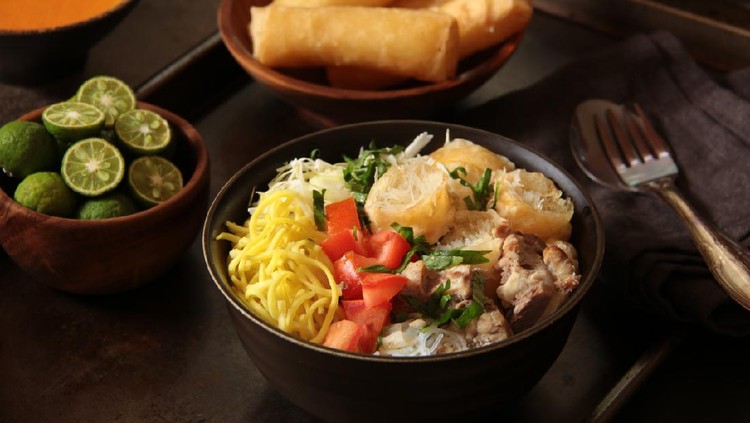 Soto Mie Bogor, the traditional Sundanese beef noodle soup. Consists of egg noodles, rice noodles, cabbage, spring roll, and tomato from Bogor, West Java. Plated in a dark colour ceramic bowl with all the toppings arranged on top of the noodles. Served with chili sauce and lime halves. Some extra spring rolls are also served along.