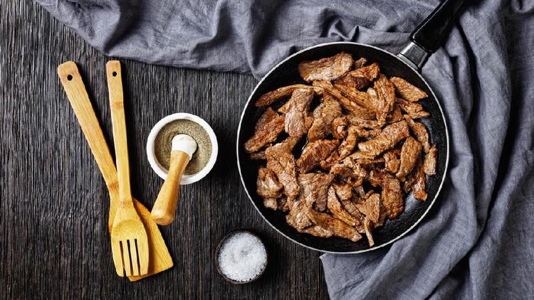 fried ribeye beef steak cut into strips on a skillet on a dark wooden table, horizontal view from above, flat lay