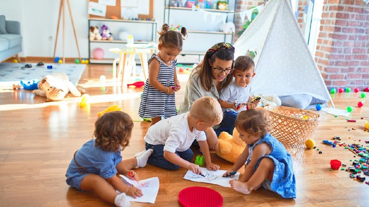 Beautiful teacher and group of toddlers sitting on the floor drawing using paper and pencil around lots of toys at kindergarten