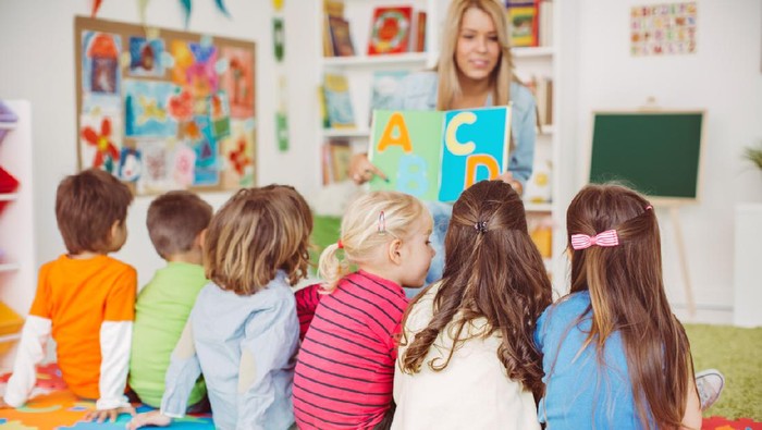 Beautiful teacher and group of toddlers sitting on the floor drawing using paper and pencil around lots of toys at kindergarten