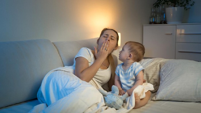 Adorable baby boy sitting on bed and looking at yawning tired mother