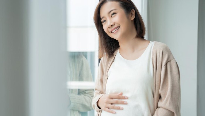 Young Asian pregnancy woman holding tummy with both hands sitting on sofa at home. Pregnant girl look, touch at stomach. Feeling of baby inside belly. Thinking of newborn infant be delivered in future