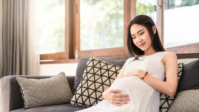 Young Asian pregnancy woman holding tummy with both hands sitting on sofa at home. Pregnant girl look, touch at stomach. Feeling of baby inside belly. Thinking of newborn infant be delivered in future