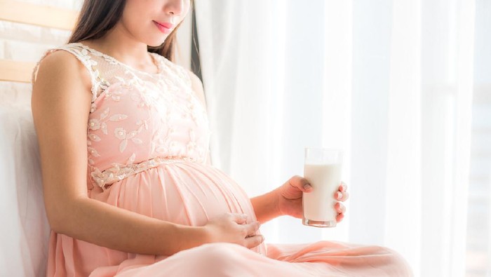 Happy pregnant woman holding a glass of fresh milk and touching her belly with care by the window in bedroom