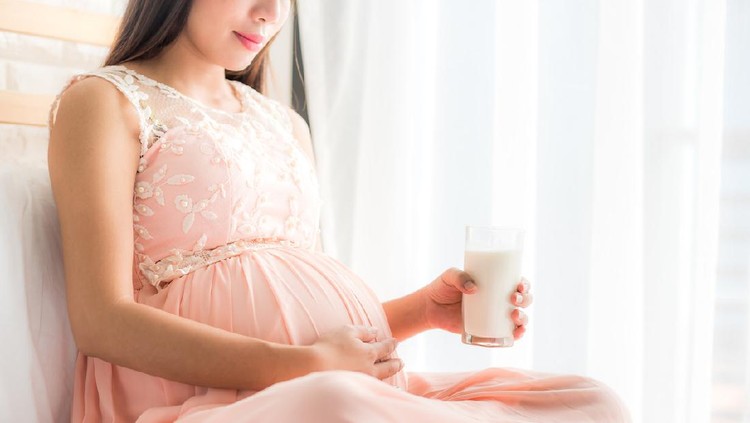 Happy pregnant woman holding a glass of fresh milk and touching her belly with care by the window in bedroom