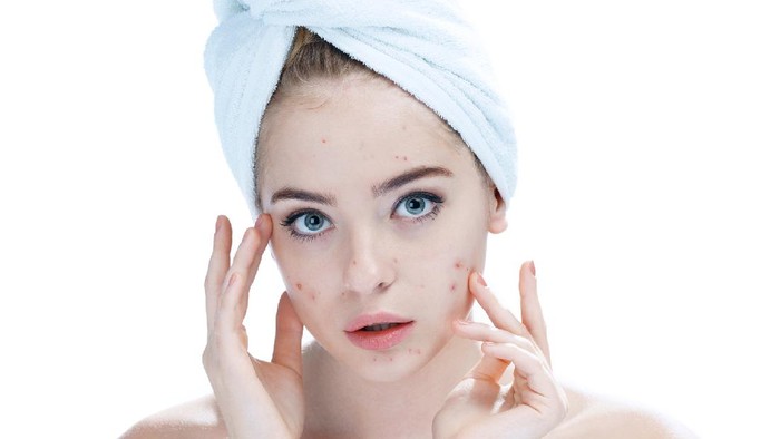 Young beautiful woman surprised to see pimple on skin. Unhappy girl with stunned funny face standing in towel in bathroom, looking in the mirror, pointing to zit