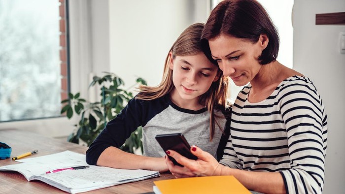 Mother using smart phone and helping daughter with homework at the table in dining room by the window