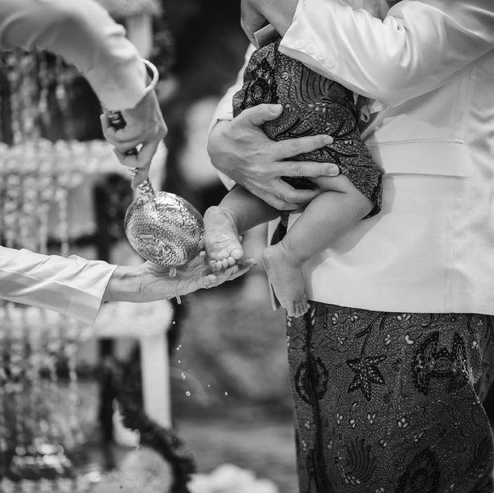 <p>Dalam foto hitam putih, Titi tampak menyiram kaki Bhaskara, Bunda. Sementara sang anak tampak nyaman digendong oleh ayahnya. (Foto: Instagram: @titiradjopadmaja)</p>