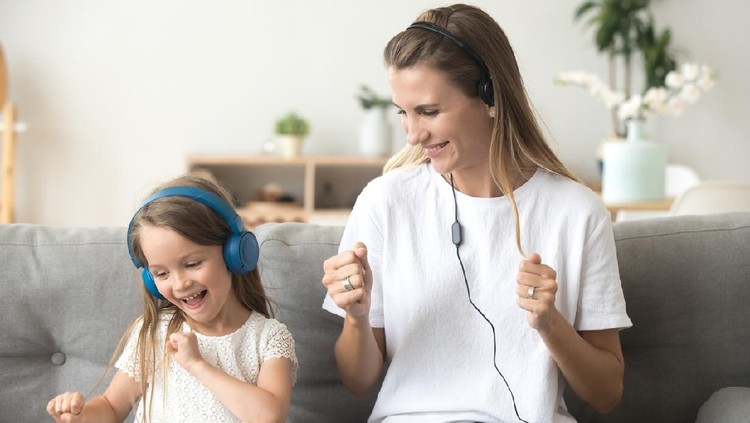 Ilustrasi Anak Dengarkan Musik Happy young mother and funny child daughter having fun laughing dancing sitting on couch listening to music in earphones together, smiling mom and kid wearing headphones enjoy favorite songs at home