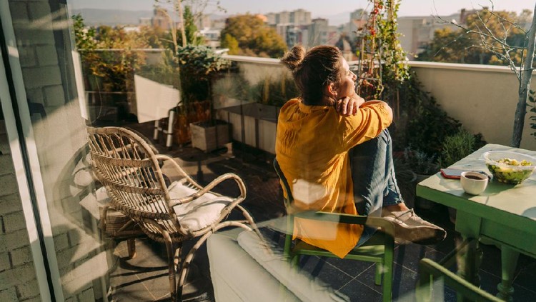 Photo of a woman drinking first morning coffee, eating 'take out' food and reading daily news online - on the balcony of her apartment while enjoying springtime sun