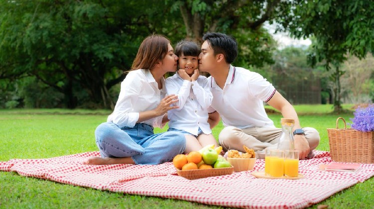 Asian teen family happy holiday picnic moment in the park with father, mother kissing daughter looking at camera and smile to happy spend vacation time together in green garden with fruit and food.