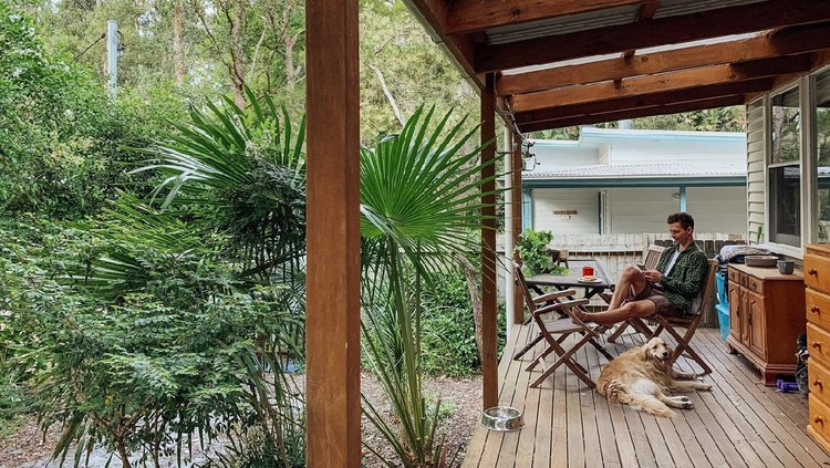 A side view shot of a young man sitting down on a chair outside his house using his smart phone with his dog.