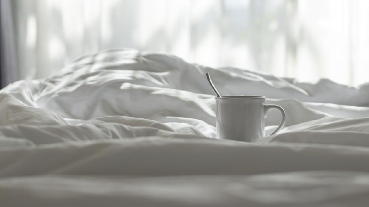 White coffee mug and spoon lay on the bed with the light from the window in the morning.