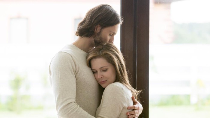 Sad young married couple embracing standing in living room opposite window at home. Sorrowful wife and husband feels unhappy, thinking about problems in relations, miscarriage, misbirth or infertility