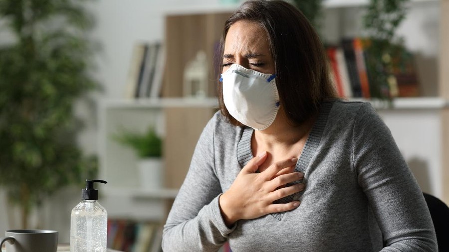 Woman with protective mask suffocating due coronavirus holding chest sitting on a desk at nigh at home