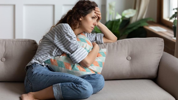 Frustrated young lady sitting on sofa, cuddling pillow, looking away at window. Lost in thoughts unhappy stressed millennial woman regretting of wrong decision, spending time alone in living room.