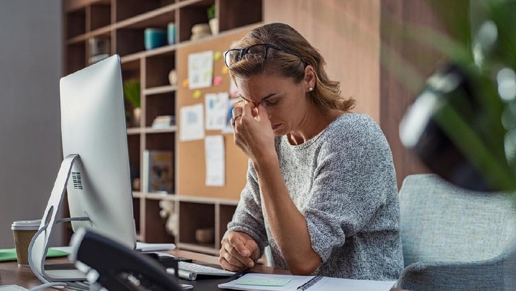 Ilustrasi Kelelahan Emosional Exhausted businesswoman having a headache in modern office. Mature creative woman working at office desk with spectacles on head feeling tired. Stressed casual business woman feeling eye pain while overworking on desktop computer.