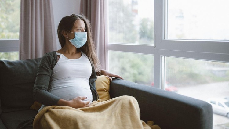 worried pregnant woman sitting at home with protective face mask and looking through the window.
