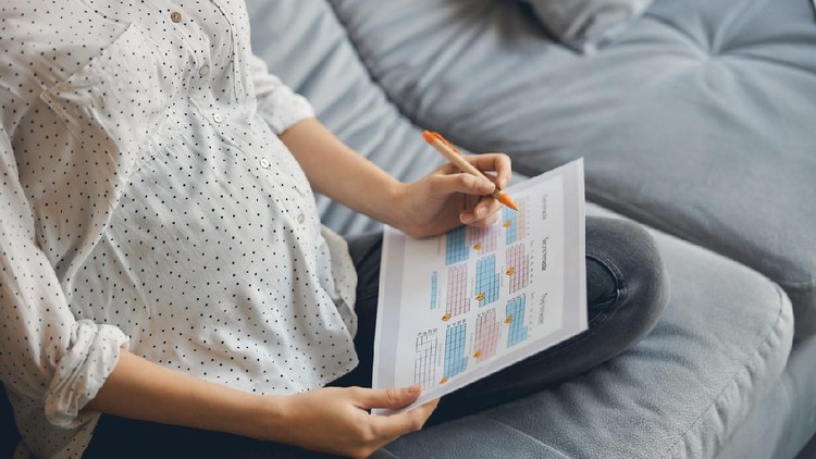 Cropped photo of a pregnant lady sitting cross legged on the sofa with a calendar in her hands and using a pen while making notes
