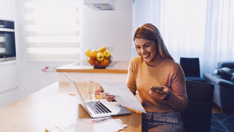 Ilustrasi wanita kerja dari rumah atau wfh atau keuangan Cute caucasian smiling blonde woman in sweater holding bills in one hand and in other smart phone. On table are laptop and bills. Apartment interior.