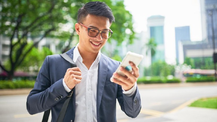 Young malaysian businessman in the streets of downtown Kuala Lumpur smiling while looking to his his smart phone for a crowed sourced taxi to arrive . Kuala Lumpur, Malaysia, Asia