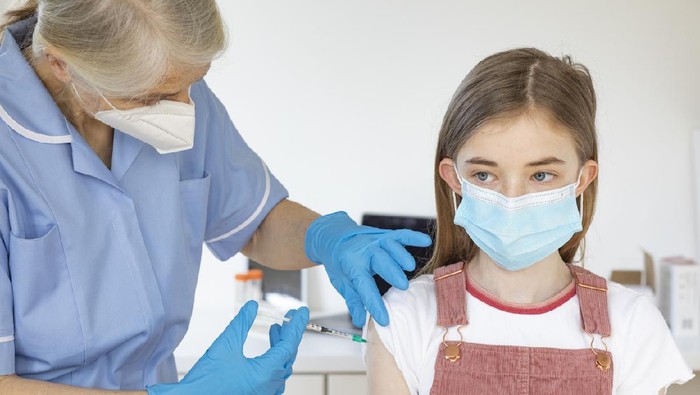 A medium shot of a Caucasian, young girl receiving her Covid-19 vaccination from a nurse. They are both wearing a protective face mask.