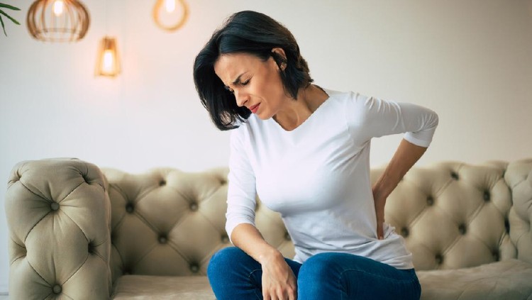 A hurting woman, who is sitting on a couch and holding her lower back with her left hand.