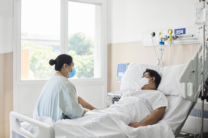 Woman visiting male patient in hospital ward. Female is sitting by man lying on bed. They are at hospital during COVID-19 epidemic. Woman visiting male patient in hospital ward. Female is sitting by man lying on bed. They are at hospital during COVID-19 epidemic.