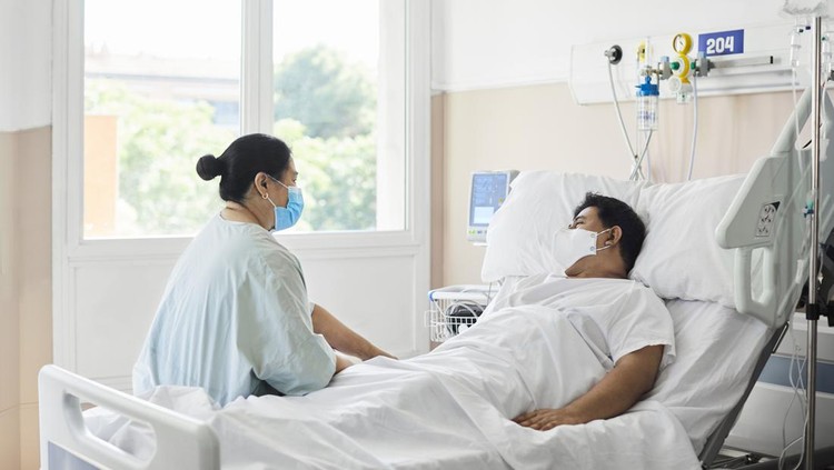 Woman visiting male patient in hospital ward. Female is sitting by man lying on bed. They are at hospital during COVID-19 epidemic.