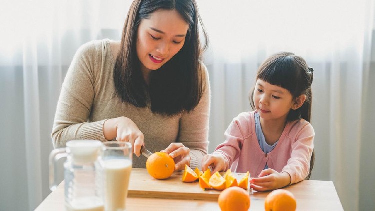 Image of Happy Young Asian mother use kitchen knife cut an orange to pieces with her daughter in living room.
